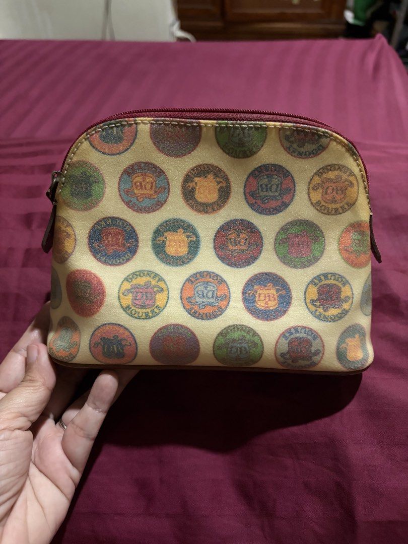Dooney & Bourke Multicolored Coin Print Cosmetic Pouch (Category: Purses & Pouches) located in Las Pinas. Submitted by ces0427 (Listing ID 1470470692, Image 1). Description: - Authentic Dooney & Bourke cosmetic pouch - Multicolored circular coin and crest print on a cream base with brown trim - Secure zip-top closure with dark red zipper fabric - Bright red fabric interior lining equipped with a convenient slip pocket - Structured base to hold it