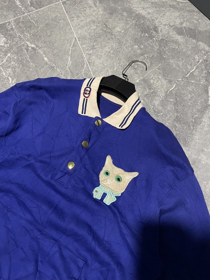 Gucci Cat Patch Blue Polo Shirt (Category: Tshirts & Polo Shirts ) located in Buenavista. Submitted by ivoperez27 (Listing ID 1470403768, Image 3). Description: - Brand: Gucci - 23 width 27 length - Color: Deep royal blue with cream and striped contrasting trims - Details: Embroidered interlocking G logo on collar, gold-tone Gucci-stamped buttons, and cute embroidered cat face patch with a light blue bow - Pockets: Front kangaroo pocket - Fit: Re