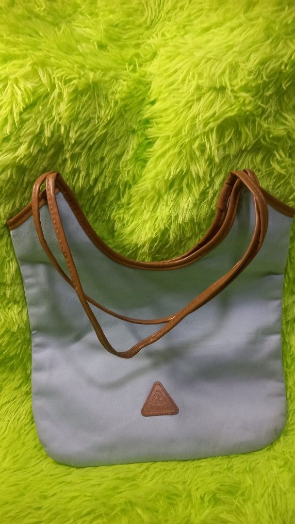 Light blue tote handbag with brown handles (Category: Tote Bags) located in Manila. Submitted by twinklelyn03 (Listing ID 1470418334, Image 2). Description: - Color: Light blue with brown faux leather trim - Design: Minimalist everyday tote with a small front triangle logo patch - Interior: Clean lining with a spacious open main compartment - Material: Lightweight fabric construction - Perfect for: Daily commute, school, running errands, or casua