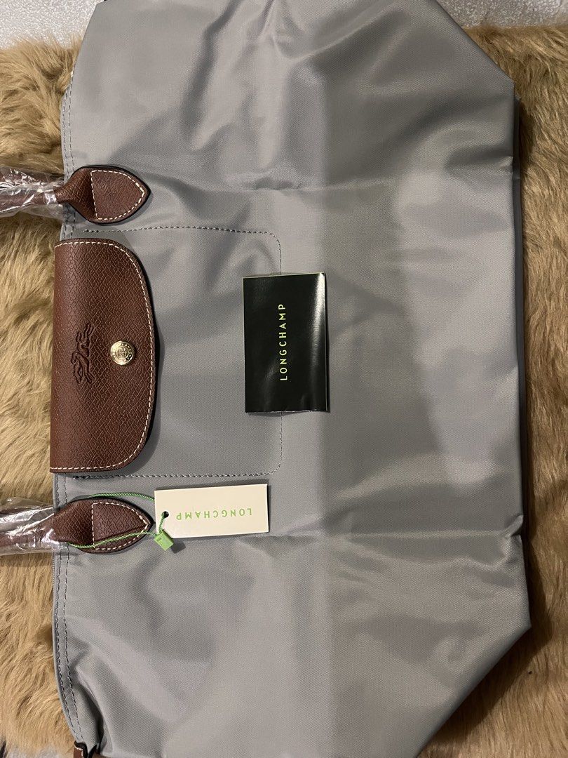 Longchamp Le Pliage Grey Tote Bag (Category: Luxury Bags & Wallets) located in Quezon City. Submitted by pickleballpaddle (Listing ID 1470528389, Image 2). Description: - Authentic Longchamp Le Pliage tote bag in a versatile grey shade - Brand new and unused condition with tags attached - Features brown leather trim, top handles, and front flap with snap closure - Gold-tone zipper pull with signature horse emblem - Handles are still wrapped in or