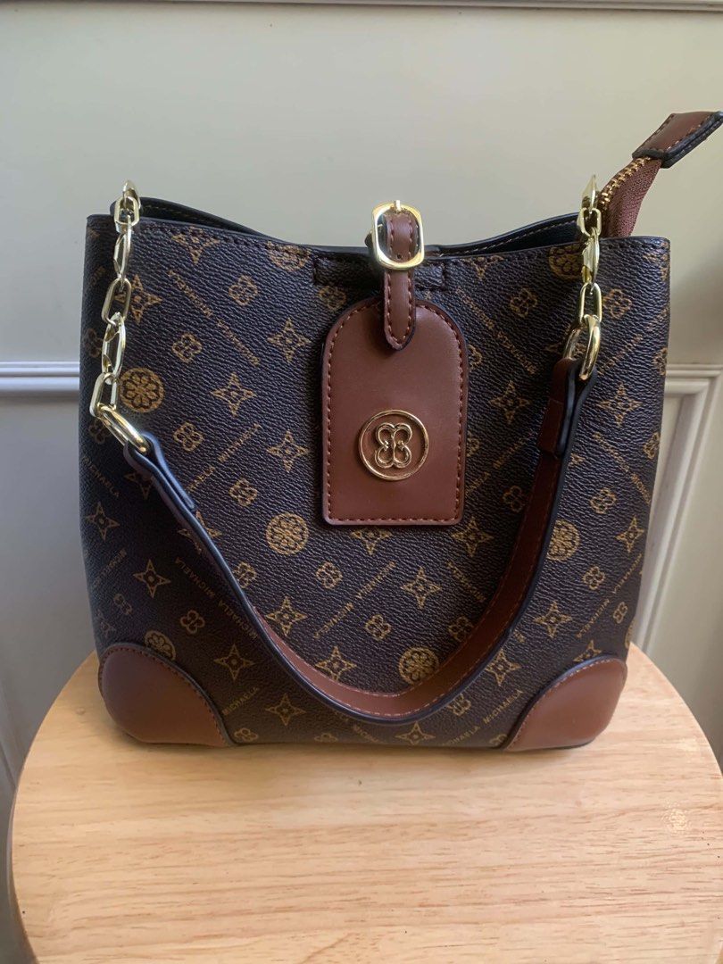 MICHAELA Monogram Bucket Bag Brown Shoulder Bag (Category: Luxury Bags & Wallets) located in Calapan. Submitted by shakalak (Listing ID 1470410815, Image 2). Description: - Brand: MICHAELA ( H 8”/L 9/W 4.5 - Style: Bucket bag in classic brown monogram print with chic tan leather trim and gold-tone hardware - Closure: Open top design with a secure strap closure - Straps: Includes a stylish chain-link shoulder strap and a detachable, adjustable lon