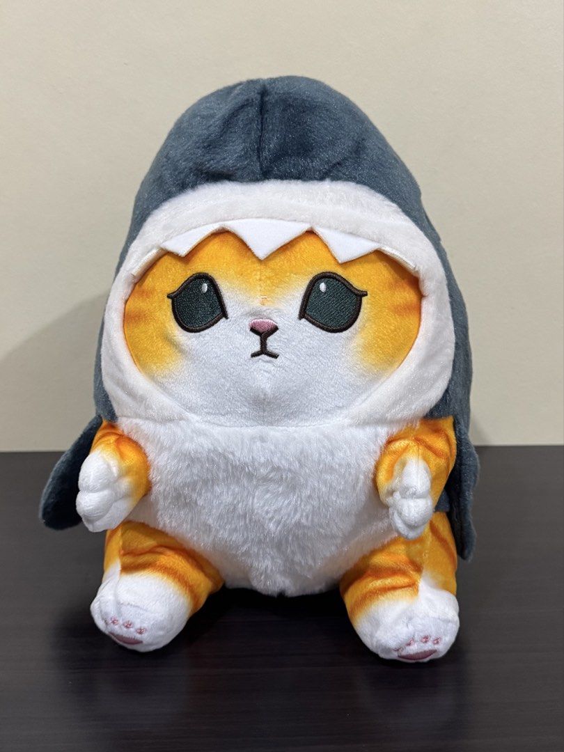 Mofusand Shark Cat Plush Stuffed Toy (Category: Plushies & Soft Toys) located in Cainta. Submitted by mayhroeen (Listing ID 1470538213, Image 1). Description: - Adorable orange tabby cat wearing a dark gray shark costume - Features white teeth framing the face and tiny embroidered paws - Made with soft fabric and fluffy filling for a cuddly feel - Includes a light blue brand tag stitched on the side - Clean condition, no stains or loose threads -