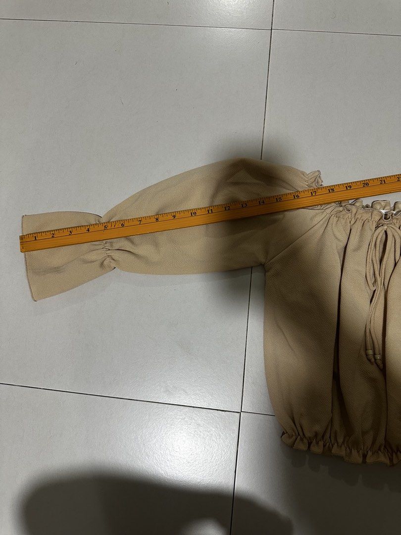 Off-shoulder beige ruffled top with tie string (Category: Blouses) located in Quezon City. Submitted by breakstuf50 (Listing ID 1470544025, Image 3). Description: Beige off-shoulder top, lightweight textured fabric. Ruffled neckline and hem with a center drawstring and wooden beads. Long bishop sleeves with flared cuffs. Measures roughly 16 inches across the chest, 15 inches down the length, and 21 inches along the sleeve. Clean and ready to wear