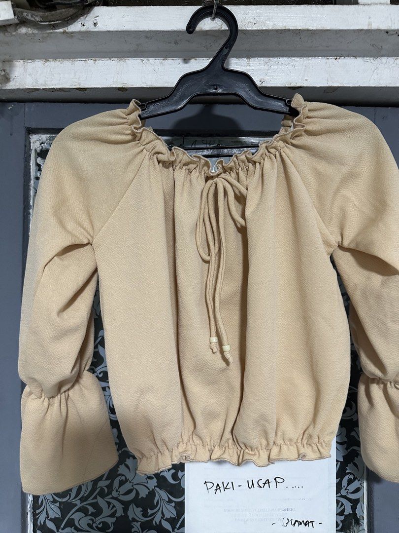 Off-shoulder beige ruffled top with tie string (Category: Blouses) located in Quezon City. Submitted by breakstuf50 (Listing ID 1470544025, Image 2). Description: Beige off-shoulder top, lightweight textured fabric. Ruffled neckline and hem with a center drawstring and wooden beads. Long bishop sleeves with flared cuffs. Measures roughly 16 inches across the chest, 15 inches down the length, and 21 inches along the sleeve. Clean and ready to wear