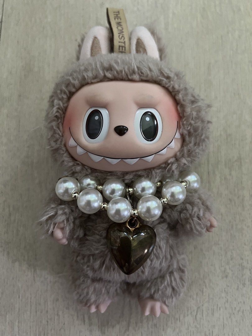 Pop Mart The Monsters Labubu Plush Doll Pendant (Category: Plushies & Soft Toys) located in Quezon City. Submitted by Zandee (Listing ID 1470488656, Image 1). Description: Pop Mart Labubu plush pendant from The Monsters series. Covered in greyish-brown fuzzy fur with soft pink ears and feet. Accessorized with a chunky double-strand pearl necklace and a dark metallic heart charm (added by owner to accesorize ) Official Pop Mart tag on top and bran