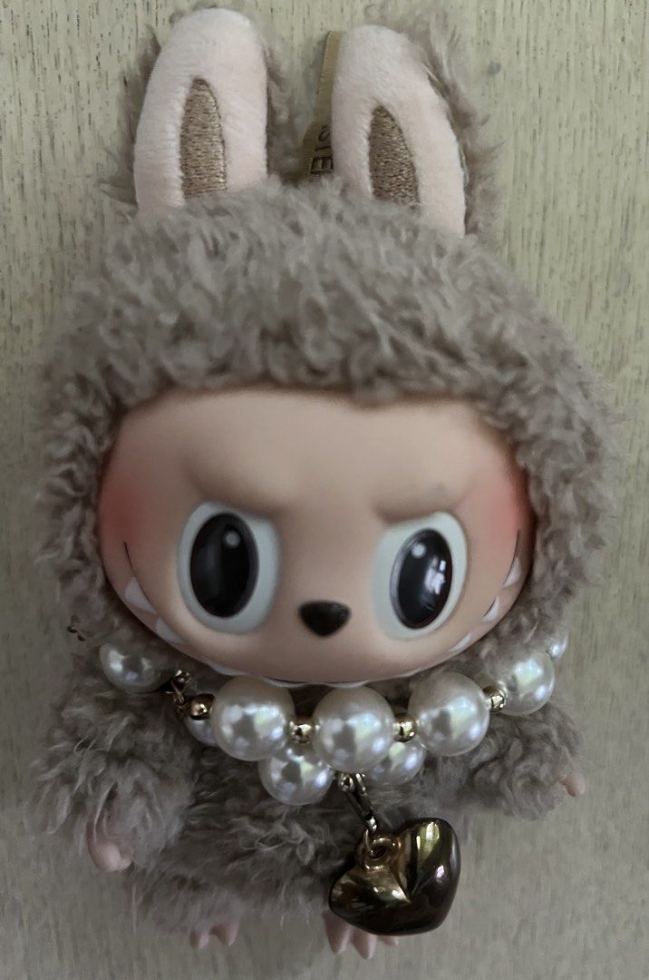 Pop Mart The Monsters Labubu Plush Doll Pendant (Category: Plushies & Soft Toys) located in Quezon City. Submitted by Zandee (Listing ID 1470488656, Image 3). Description: Pop Mart Labubu plush pendant from The Monsters series. Covered in greyish-brown fuzzy fur with soft pink ears and feet. Accessorized with a chunky double-strand pearl necklace and a dark metallic heart charm (added by owner to accesorize ) Official Pop Mart tag on top and bran