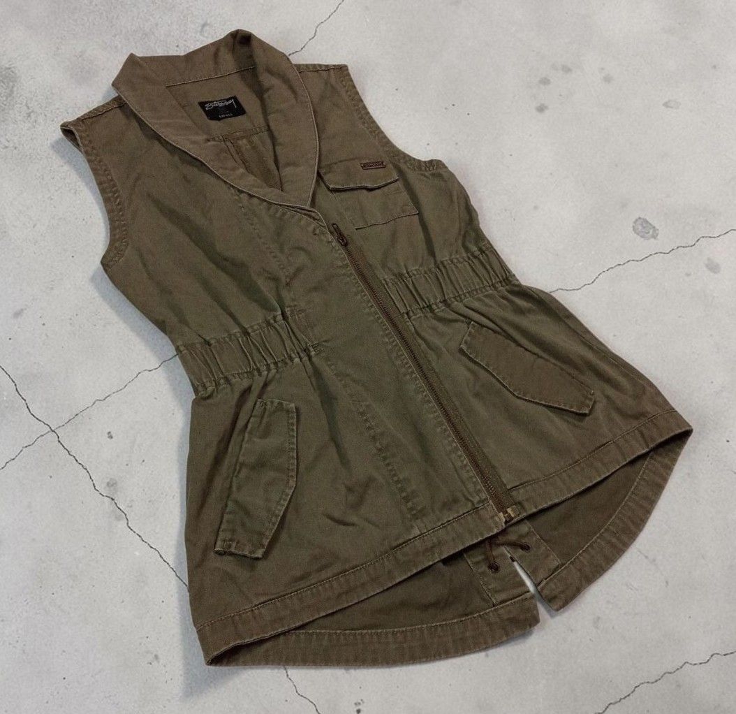 Stussy Olive Green Utility Vest (Category: Blouses) located in Manila. Submitted by grmnts_frmthe_slmz (Listing ID 1470391103, Image 2). Description: Olive green Stussy vest, size small. Cotton fabric with a textured vertical weave. Asymmetric front zipper closure and structured collar. Elasticized waist cinches the fit nicely. Multiple utility pockets, including one chest pocket with a metal Stussy logo badge. Curved hem at the back with drawstr