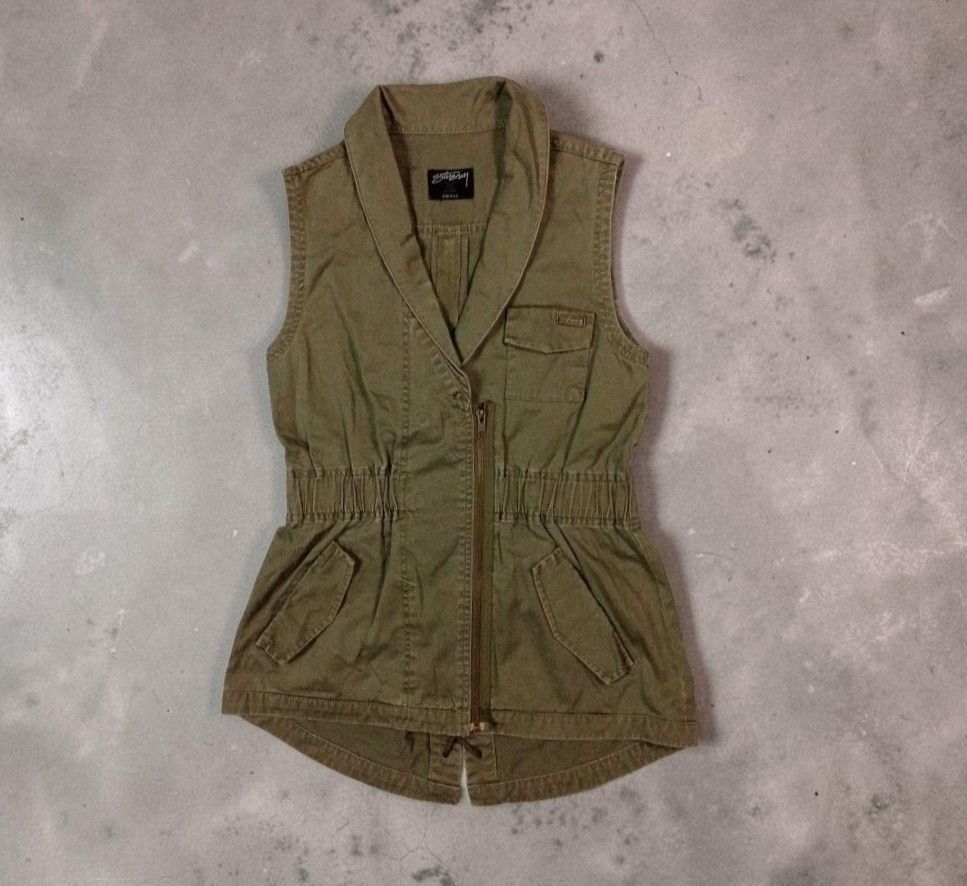 Stussy Olive Green Utility Vest (Category: Blouses) located in Manila. Submitted by grmnts_frmthe_slmz (Listing ID 1470391103, Image 1). Description: Olive green Stussy vest, size small. Cotton fabric with a textured vertical weave. Asymmetric front zipper closure and structured collar. Elasticized waist cinches the fit nicely. Multiple utility pockets, including one chest pocket with a metal Stussy logo badge. Curved hem at the back with drawstr