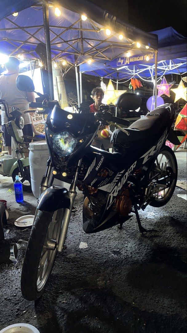 Suzuki Raider R150 Underbone Motorcycle (Category: Motorbikes for Sale) located in Manila. Submitted by miksnmatchs (Listing ID 1470556262, Image 1). Description: Suzuki Raider R150 in black with white graphics. Standard spoke wheels, custom orange engine crankcase cover, aftermarket exhaust pipe installed. Dual front lights mounted near the forks. Standard single seat, black frame, manual chain drive setup. Registered plate visible at the rear. 