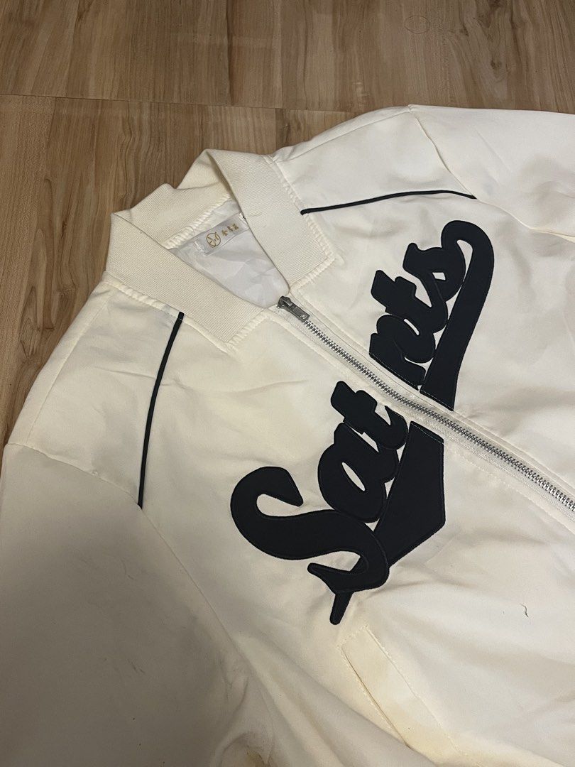 White Saints Bomber Jacket (Category: Coats, Jackets and Outerwear) located in San Fernando. Submitted by angelhave_wings (Listing ID 1470552081, Image 2). Description: - Color: White with bold black 'Saints' script across the chest - Fit: Regular fit, lightweight fabric ideal for layering - Design: Zip-up front with stylish black and white striped ribbing on collar, cuffs, and hem - Extra Feature: Functional utility zip pocket on the left sleeve