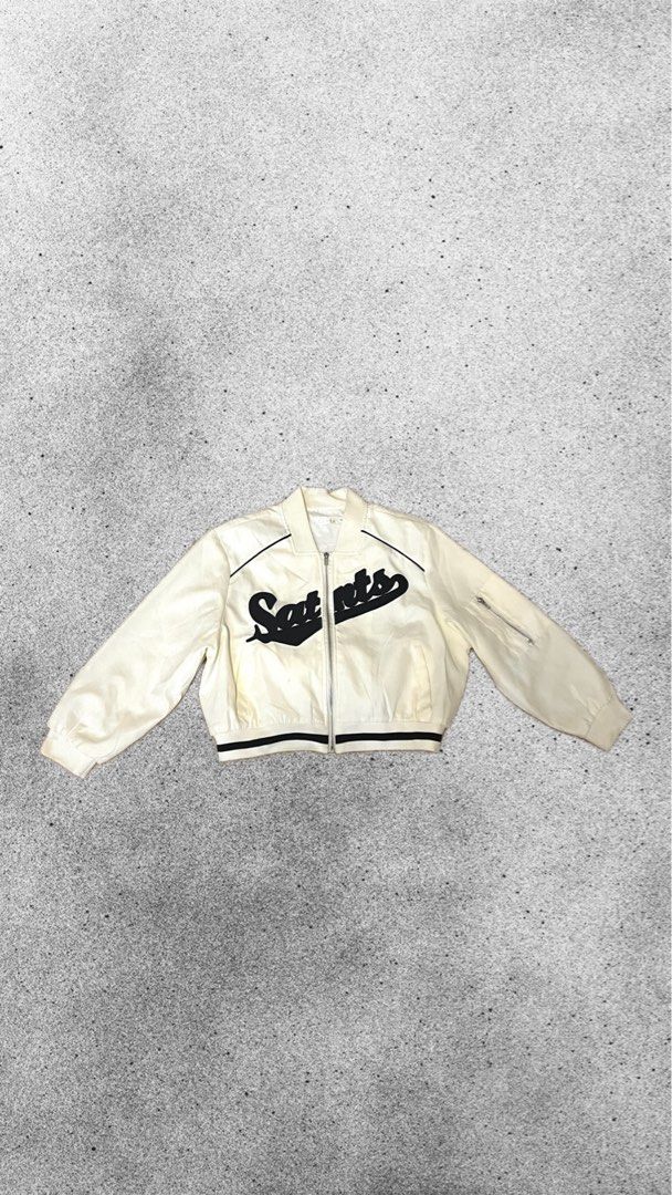 White Saints Bomber Jacket (Category: Coats, Jackets and Outerwear) located in San Fernando. Submitted by angelhave_wings (Listing ID 1470552081, Image 1). Description: - Color: White with bold black 'Saints' script across the chest - Fit: Regular fit, lightweight fabric ideal for layering - Design: Zip-up front with stylish black and white striped ribbing on collar, cuffs, and hem - Extra Feature: Functional utility zip pocket on the left sleeve