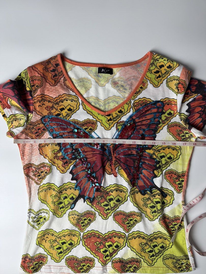 Y2K Butterfly Sequin Graphic V-Neck Tee (Category: Shirts) located in Manila. Submitted by babygirly2k (Listing ID 1470403223, Image 3). Description: - Style: Y2K short-sleeve top - Design: White base with all-over colorful heart and butterfly pattern - Details: Large embroidered front butterfly with blue glitter edging and red sequins - Trim: Orange ribbed V-neckline - Branding: 'Ken' label on inside collar - Measurements: 17" pit-to-pit, 22" le