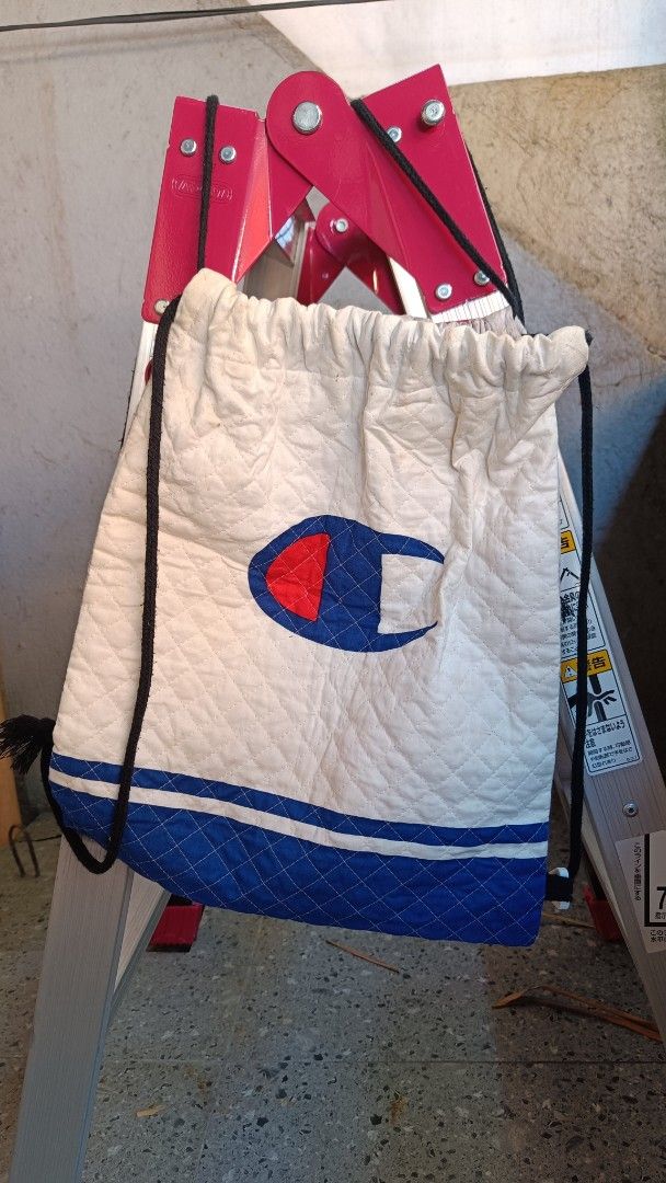 💯 Original Champion Quilted Drawstring Backpack Bag (Category: Backpacks) located in Cavite. Submitted by randzwurkz48441 (Listing ID 1470630559, Image 2). Description: - Brand: Champion - Dimensions: 15.5 x 12.5 inches - Type: Quilted drawstring bag - Color: Light Grey body with blue bottom panel and white accent stripes - Details: Classic red and blue Champion logo on the front - Straps: Durable black rope cords for shoulder wear - Condition: 