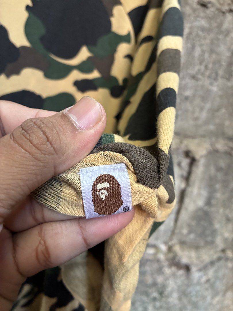 A Bathing Ape Bape Camo T-Shirt (Category: Tshirts & Polo Shirts ) located in Manila. Submitted by assortedgoodsv3 (Listing ID 1470620963, Image 3). Description: - Brand: A Bathing Ape (BAPE) - Design: Yellow camouflage all-over print (brown, green, and black tones) - Fit: Relaxed, casual streetwear - Details: Short sleeves, classic crew neck, iconic ape head logo tag near collar - Condition: Soft and worn-in fabric, ready for daily wear.