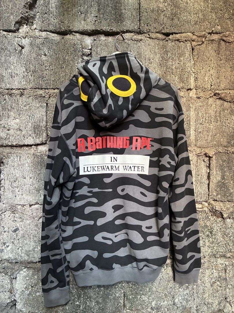 A Bathing Ape Camo Zip Up Hoodie (Category: Hoodies) located in Manila. Submitted by assortedgoodsv3 (Listing ID 1470619950, Image 3). Description: - Brand: A Bathing Ape (BAPE) - Design: Black and grey camo pattern with yellow hood graphics - Details: Ring pull zipper and front pockets - Back Graphic: Red and white text block lettering reading "A Bathing Ape in Lukewarm Water" - Fit: Ribbed cuffs and hem for comfort - Condition: Excellent, ready
