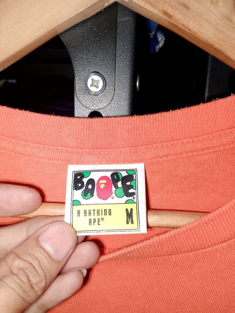 A Bathing Ape Shark Face Orange T-Shirt (Category: Tshirts & Polo Shirts ) located in Manila. Submitted by mjthrifted_second (Listing ID 1470614575, Image 2). Description: - Brand: A Bathing Ape (BAPE) - Color: Orange - Size: Medium -Dimes: 21/27 - Design: Small embroidered shark graphic on chest - Details: Classic sleeve tag near hem, printed neck label - Condition: Clean fabric, ready to wear.