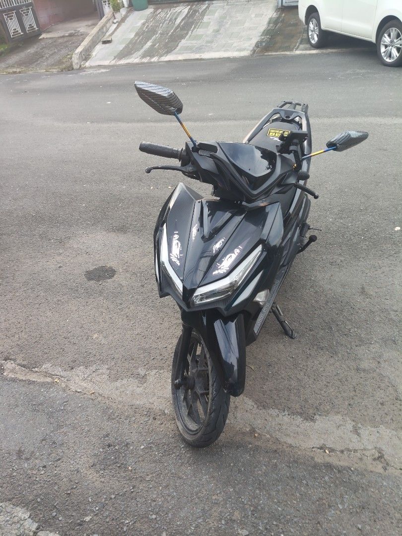 Black Scooter Motorcycle (Category: Motorbikes for Sale) located in Manila. Submitted by zealphoenix.c08cc4 (Listing ID 1470721637, Image 2). Description: Black scooter parked outside on asphalt. Features carbon-pattern mirrors, phone mount on handlebars, and side kickstand down. Body has some white graphic stickers near the front headlight. Rear cargo rack installed behind seat. Rear tire tread visible with brake lever and kick start pedal near 
