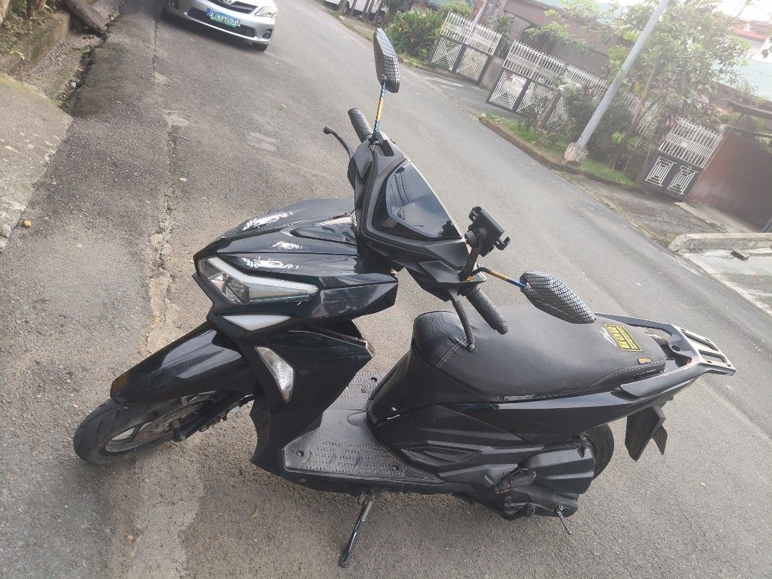 Black Scooter Motorcycle (Category: Motorbikes for Sale) located in Manila. Submitted by zealphoenix.c08cc4 (Listing ID 1470721637, Image 3). Description: Black scooter parked outside on asphalt. Features carbon-pattern mirrors, phone mount on handlebars, and side kickstand down. Body has some white graphic stickers near the front headlight. Rear cargo rack installed behind seat. Rear tire tread visible with brake lever and kick start pedal near 