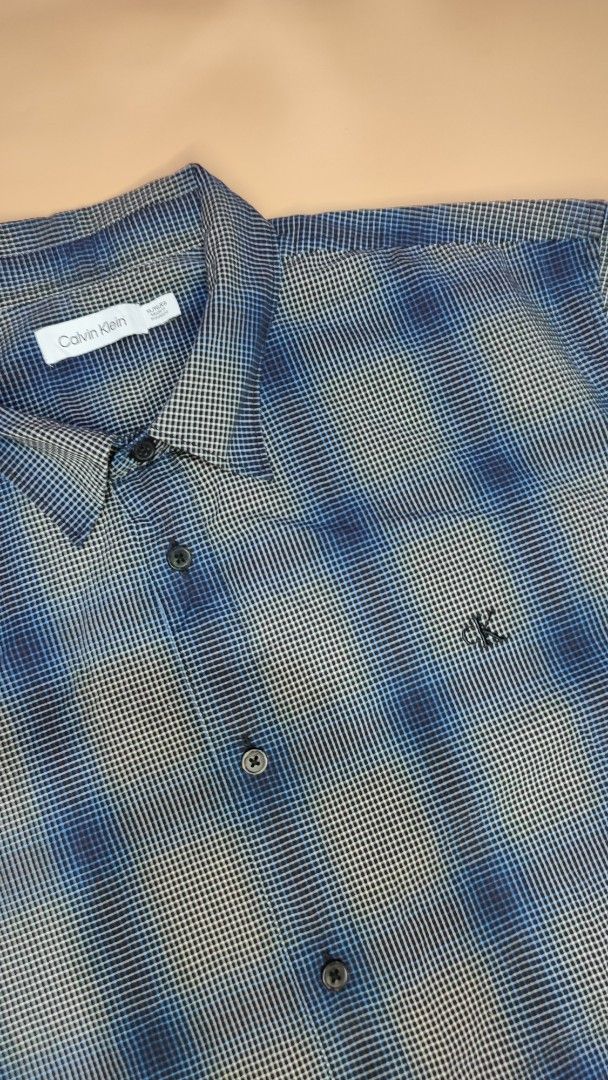 Calvin Klein Blue Checked Short Sleeve Shirt (Category: Tshirts & Polo Shirts ) located in Cavite. Submitted by juanpraktikal (Listing ID 1470738112, Image 2). Description: - Brand: Calvin Klein - Size: XL 25" x 30.5" Length - Color/Pattern: Blue and grey checked - Material: 97% cotton, 3% elastane blend (slight stretch) - Details: Regular button-down closure, dark buttons, classic collar, chest pocket with small embroidered ck logo - Condition: 