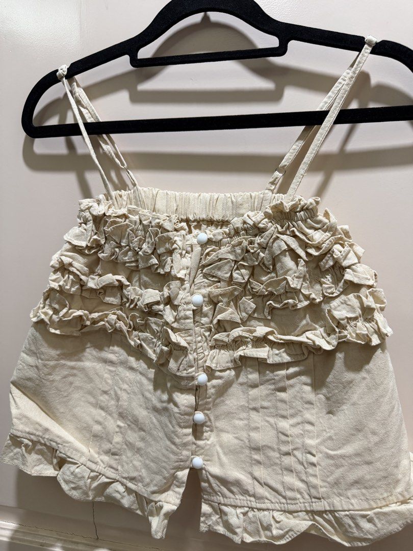 Cream Ruffle Cami Top with Buttons (Category: Sleeveless) located in Davao. Submitted by 2ndbase.dvo.ii (Listing ID 1470596297, Image 3). Description: - Color: Cream - Style: Cottagecore / Coquette - Details: Ruffled chest and hem, pleated lower front, round white buttons - Straps: Adjustable thin shoulder straps - Closure: Button-down front - Fabric: Light and breathable, ideal for warmer weather - Condition: Excellent, no major flaws noticed.