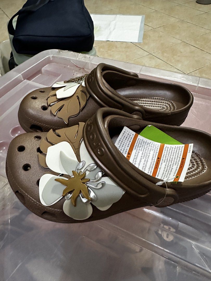 Crocs Brown Floral Clogs (Category: Slippers and slides) located in Balanga. Submitted by jamilbeauty5 (Listing ID 1470597950, Image 3). Description: - Brand new brown Crocs clogs in slip-on style - Decorated with trendy white floral and monstera leaf charms - Features ventilation holes on the toe box for breathability - Adjustable brown pivoting heel strap with Crocs branding - Comes with original product tag and plastic protective wrapping - Gu