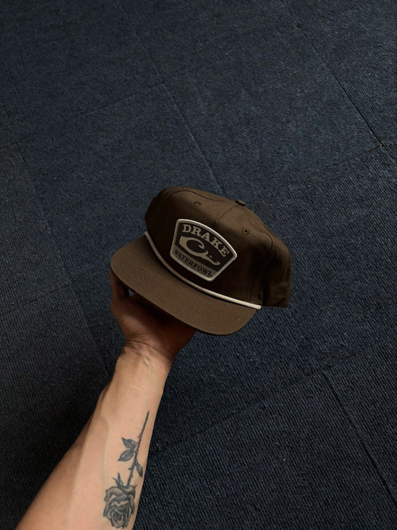Drake Waterfowl Brown Snapback Cap (Category: Caps & Hats) located in Manila. Submitted by toys_hats005 (Listing ID 1470629528, Image 1). Description: - Brand/Style: Drake Waterfowl snapback hat in brown - Design: Structured crown, flat brim, and stylish contrast rope detail across the front - Details: Front patch logo and Lost brand tab at the back snap - Comfort: Clean inside lining with custom branded taping - Condition/Fit: Adjustable snap cl