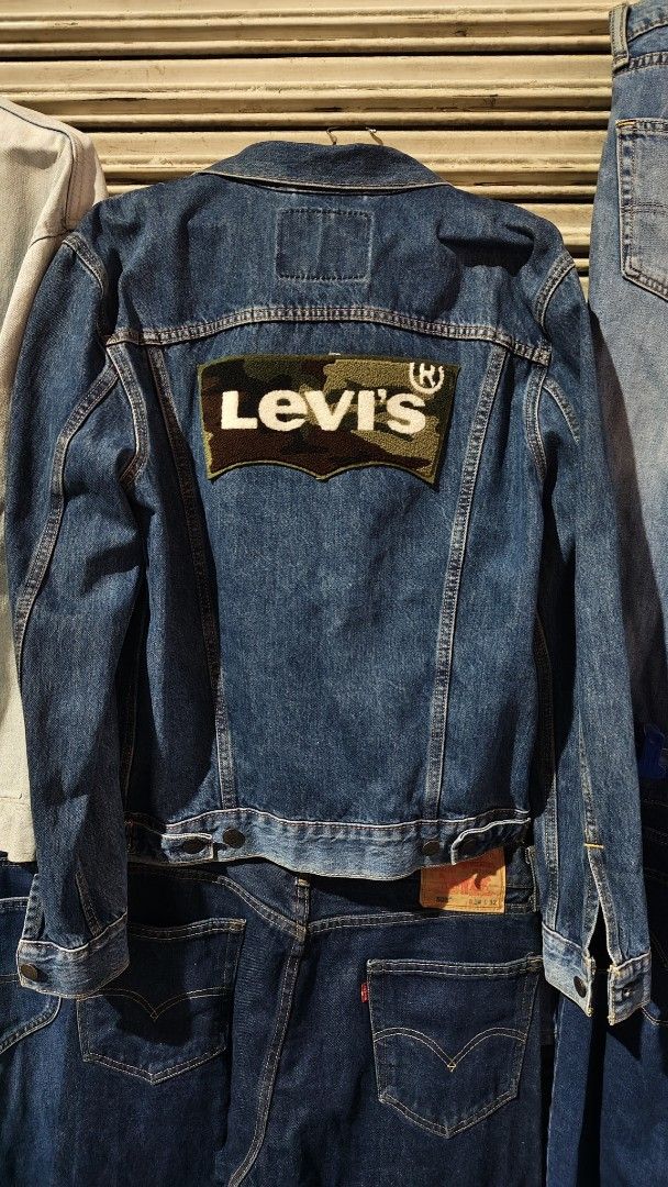 Levi's Denim Trucker Jacket Size M (Category: Coats, Jackets and Outerwear) located in Quezon City. Submitted by truedeer_cda94c (Listing ID 1470606011, Image 2). Description: - Brand: Levi's - Style: Denim jacket with classic button-up front, chest pockets, and side pockets - Color/Wash: Medium blue wash - Size: Medium (M) - Special Feature: Textured camouflage Levi's logo patch stitched across the back - Details: Includes interior wash tags and