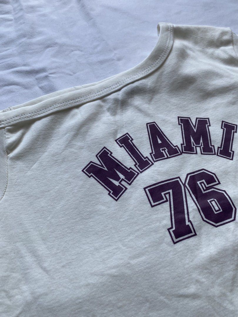 Miami 76 Asymmetrical White Crop Top (Category: Shirts) located in Valenzuela. Submitted by mandies_25 (Listing ID 1470667760, Image 2). Description: - Style: Asymmetrical one-shoulder crop top with short sleeves - Design: Purple 'Miami 76' varsity graphic on the chest - Fit: Snug fit, stretchy cotton fabric for comfort - Condition: Clean, no stains or holes (wardrobe clear-out) - Easy to pair with high-waisted jeans or skirts - Small.