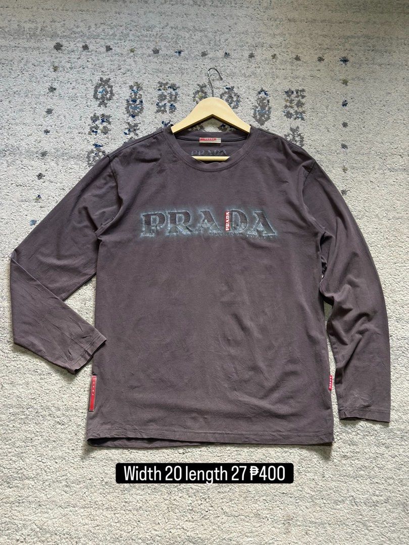 Prada Grey Long Sleeve T-Shirt (Category: Longsleeves) located in Daet. Submitted by thrifthings2018 (Listing ID 1470618422, Image 1). Description: - Brand: Prada - Item: Long sleeve tee - Color: Dark grey - Material: 100% cotton - Design: Bold distressed Prada logo across the chest with signature red Prada tag on hem and inner collar - Measurements: Width 20 inches, Length 27 inches - Care: Machine wash warm - Condition: Excellent / Well-maintai