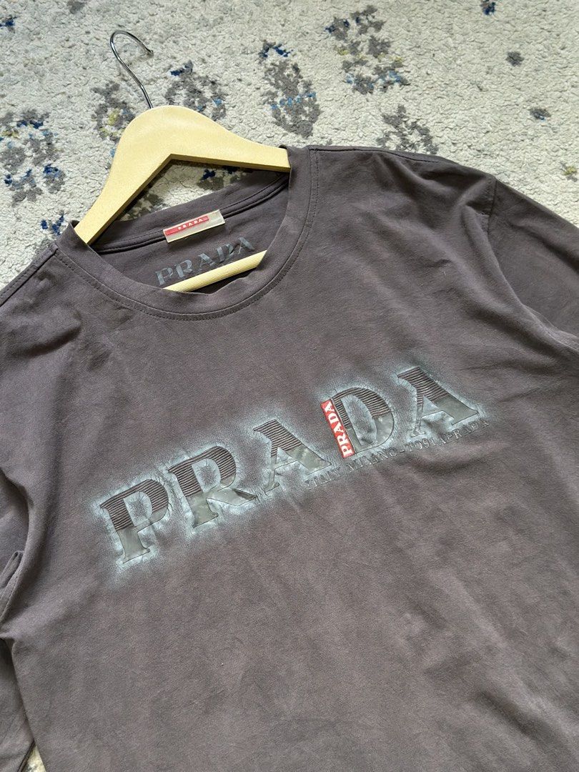 Prada Grey Long Sleeve T-Shirt (Category: Longsleeves) located in Daet. Submitted by thrifthings2018 (Listing ID 1470618422, Image 2). Description: - Brand: Prada - Item: Long sleeve tee - Color: Dark grey - Material: 100% cotton - Design: Bold distressed Prada logo across the chest with signature red Prada tag on hem and inner collar - Measurements: Width 20 inches, Length 27 inches - Care: Machine wash warm - Condition: Excellent / Well-maintai