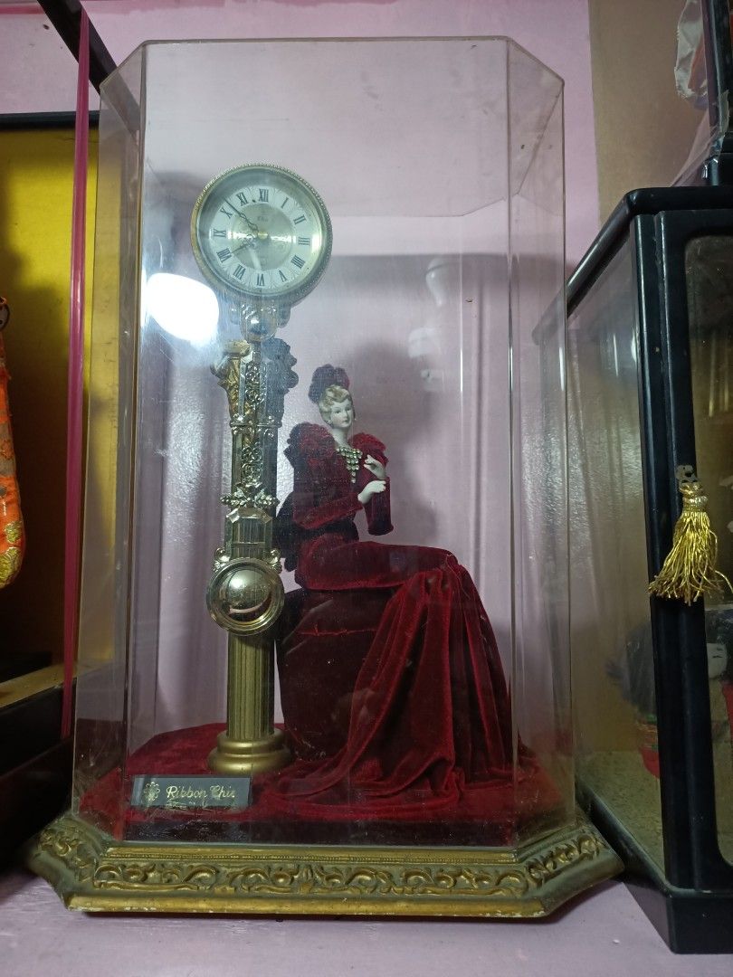 Ribbon Chic Figurine Clock with Glass Case (Category: Clocks) located in Taguig. Submitted by grandfish_55ab5c (Listing ID 1470568930, Image 1). Description: - Elegant decorative clock with a seated female figurine in a striking red velvet dress - Encased in a stylish tall hexagonal glass display case with a luxurious gold-toned base and trim - Features a classic Roman numeral clock face mounted on an ornate pillar structure - Perfect unique coll