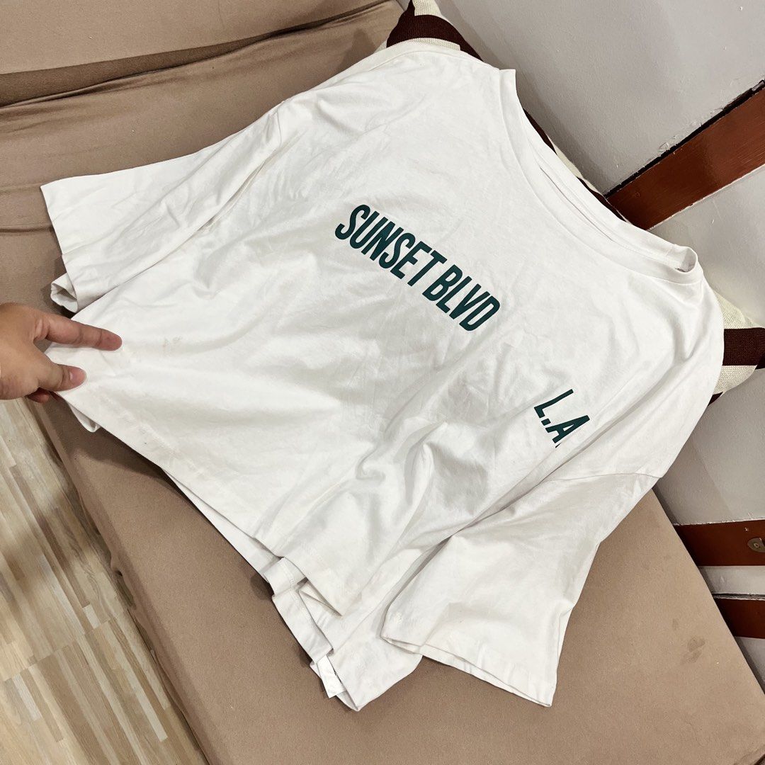 Sunset Blvd L.A. White Graphic Tee (Category: Tshirts & Polo Shirts ) located in Manila. Submitted by mjsappstopandshop (Listing ID 1470695525, Image 1). Description: White oversized t-shirt with dark green graphic print. Text reads on the front and back, with California Los Angeles lettering and star graphics below. Short sleeves, relaxed fit, comfy cotton material. Great casual piece for everyday wear..
