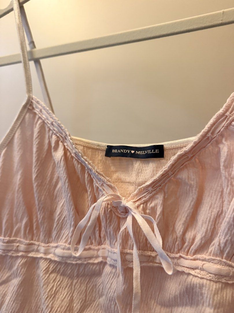 Brandy Melville Light Pink Cami Tank Top (Category: Sleeveless) located in Taguig. Submitted by rleilhoreine (Listing ID 1470769811, Image 2). Description: - Brand: Brandy Melville - Color: Pink - Style: Cami top with sweetheart neckline, front tie bow, and lettuce hem - Features: Adjustable straps and textured crinkle fabric - Condition: Good overall shape with small stains on the back near the top seam .