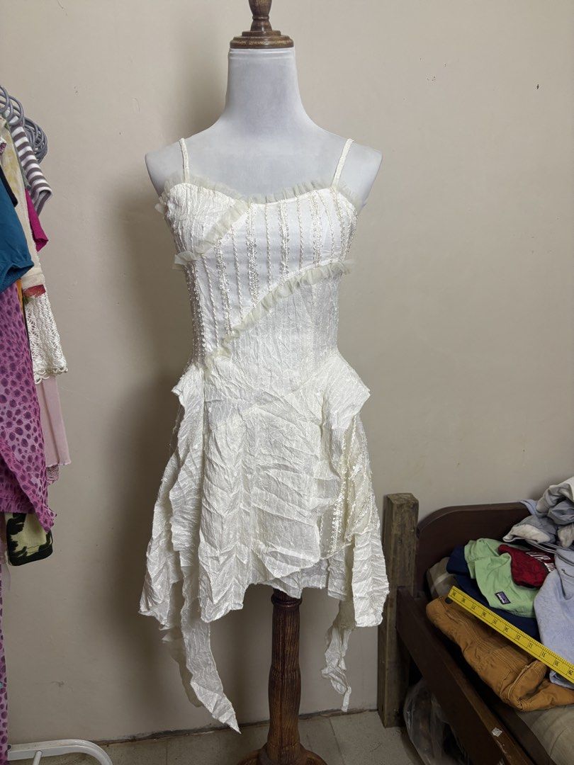 fairycore/Y2K-style camisole mini dress (Category: Dresses) located in Ajuy. Submitted by galitsabarat (Listing ID 1470793320, Image 1). Description: - Color: White - Style: Sleeveless with delicate spaghetti straps - Bodice: Structured corset style with a stylish diagonal ruffle trim - Fabric: Premium textured material throughout - Waistline: Trendy drop-waist design - Skirt: Layered, asymmetrical handkerchief hem - Closure: Secure back zipper -