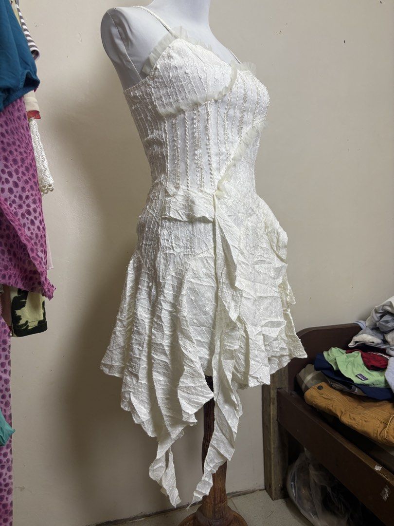 fairycore/Y2K-style camisole mini dress (Category: Dresses) located in Ajuy. Submitted by galitsabarat (Listing ID 1470793320, Image 3). Description: - Color: White - Style: Sleeveless with delicate spaghetti straps - Bodice: Structured corset style with a stylish diagonal ruffle trim - Fabric: Premium textured material throughout - Waistline: Trendy drop-waist design - Skirt: Layered, asymmetrical handkerchief hem - Closure: Secure back zipper -