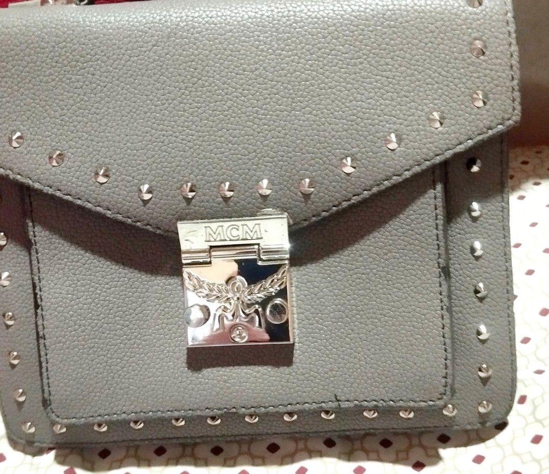 MCM Grey Studded Leather Crossbody Bag (Category: Cross-body Bags) located in Manila. Submitted by lene48 (Listing ID 1470853754, Image 1). Description: - Brand: MCM - Material: Grey pebbled leather with silver-tone hardware and stud details - Closure: Push-lock closure with engraved brand logo - Interior: Vibrant red lining featuring a zipper pocket and metal brand plaque - Straps: Structured top handle and removable shoulder strap for versatile