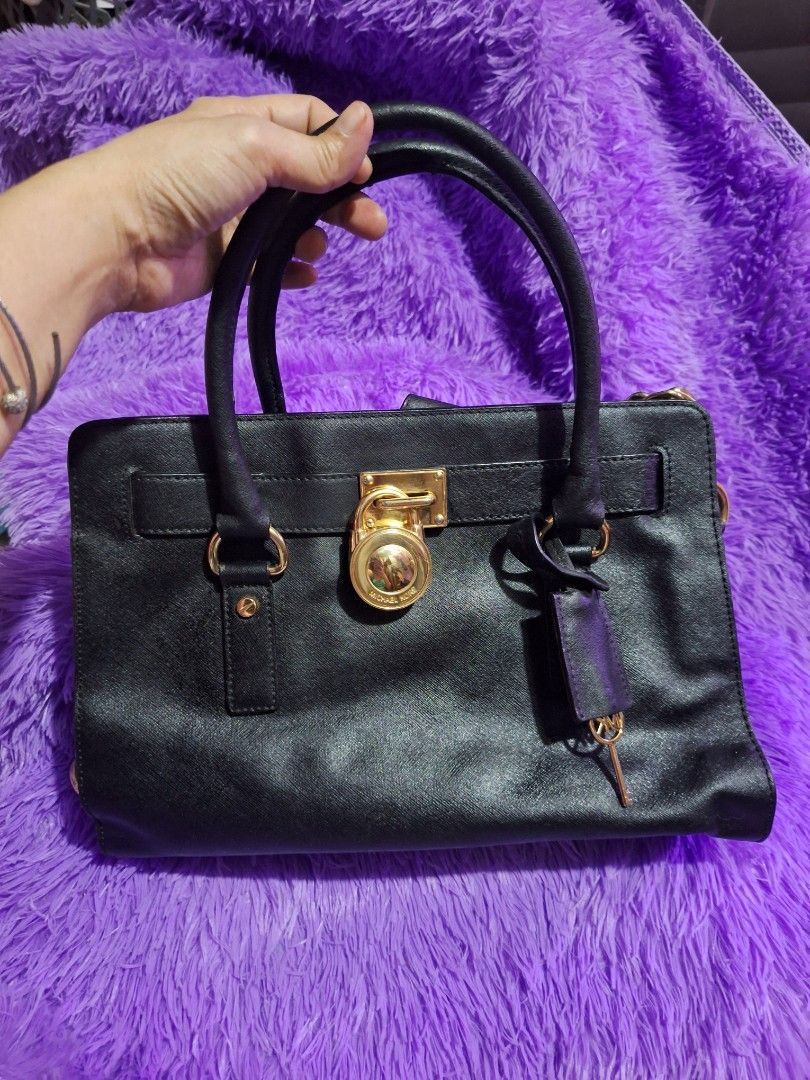 Michael Kors Black Hamilton Handbag With Gold Hardware (Category: Luxury Bags & Wallets) located in Quezon City. Submitted by cloudymay (Listing ID 1470820362, Image 1). Description: Black Michael Kors satchel bag. Gold lock detail on the front, plus a chunky gold chain strap on the side. Double top handles. Tan fabric lining inside with zipper pockets. Structured shape with pebbled texture. Zip closure on top. Min signs of wear on leather: folds