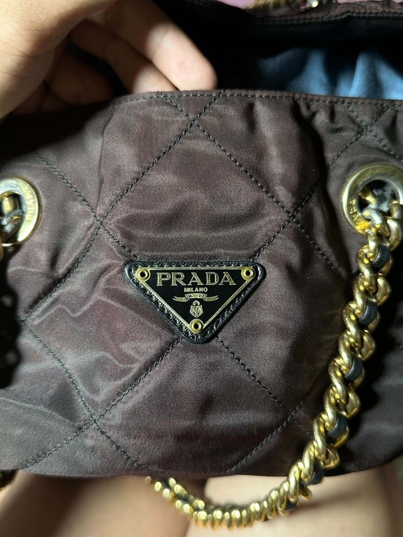 Prada Quilted Nylon Chain Tote Bag Brown (Category: Luxury Bags & Wallets) located in Naga. Submitted by mrwisethrifter (Listing ID 1470853957, Image 2). Description: - Brand: Prada - Material: Quilted nylon with Saffiano leather trim - Color: Brown with gold-tone hardware - Straps: Gold-tone chain interwoven with leather - Hardware: Iconic triangular Prada logo plaque on the front - Closure: Saffiano leather snap strap at the top - Interior: Bla
