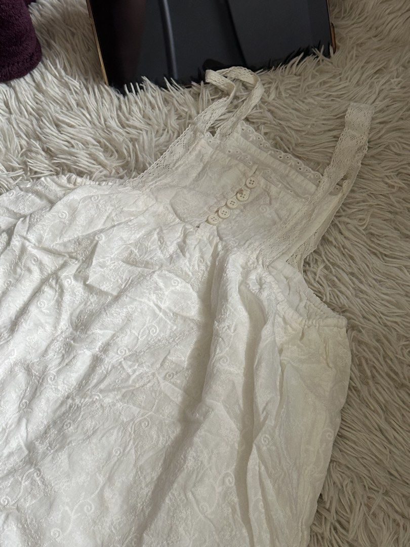 White lace trim tank top with buttons (Category: Sleeveless) located in Taguig. Submitted by appatheseller (Listing ID 1470845686, Image 2). Description: Small stain on chest area not visible - Color: White - Style: Sleeveless top with delicate lace trim along the neckline and straps - Details: Front row of small white buttons, textured fabric with subtle pattern - Fit: Elastic gathering under the chest area for a flattering silhouette - Material