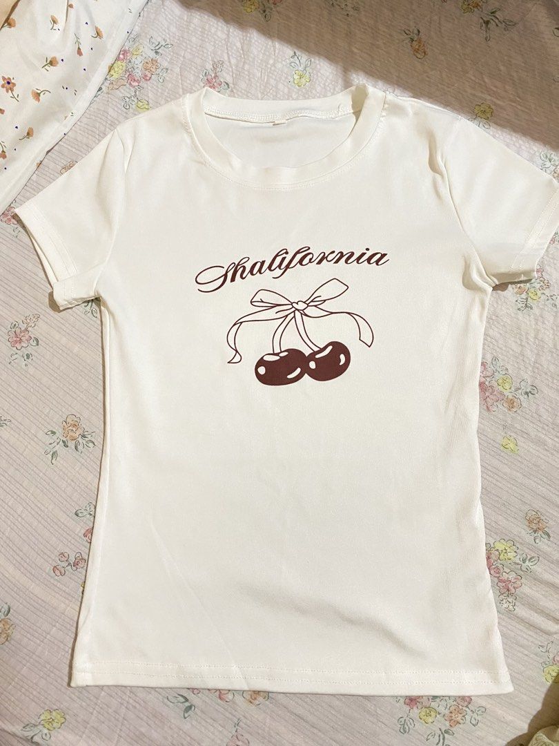 White T-Shirt with Cherry Print and Bow (Category: Shirts) located in Imus. Submitted by softclosetph (Listing ID 1470920339, Image 3). Description: - Style: Y2K fitted short-sleeve tee with crew neckline - Design: 'Shalifornia' text with cute brown cherry and bow graphic - Condition: Clean fabric, no stains or flaws, ready to wear - Pairing: Looks great with high-waisted jeans or skirts.