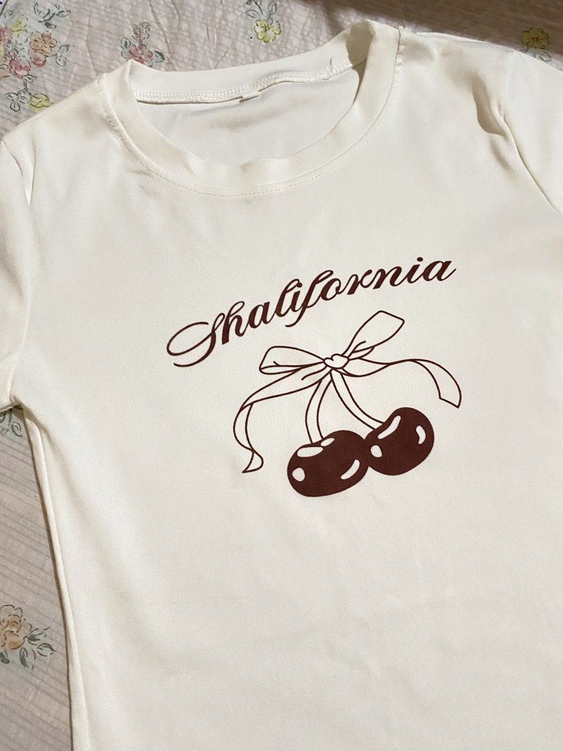 White T-Shirt with Cherry Print and Bow (Category: Shirts) located in Imus. Submitted by softclosetph (Listing ID 1470920339, Image 2). Description: - Style: Y2K fitted short-sleeve tee with crew neckline - Design: 'Shalifornia' text with cute brown cherry and bow graphic - Condition: Clean fabric, no stains or flaws, ready to wear - Pairing: Looks great with high-waisted jeans or skirts.