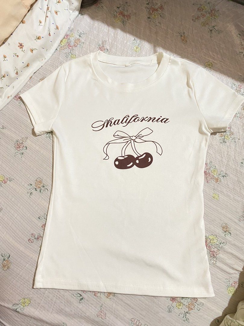 White T-Shirt with Cherry Print and Bow (Category: Shirts) located in Imus. Submitted by softclosetph (Listing ID 1470920339, Image 1). Description: - Style: Y2K fitted short-sleeve tee with crew neckline - Design: 'Shalifornia' text with cute brown cherry and bow graphic - Condition: Clean fabric, no stains or flaws, ready to wear - Pairing: Looks great with high-waisted jeans or skirts.