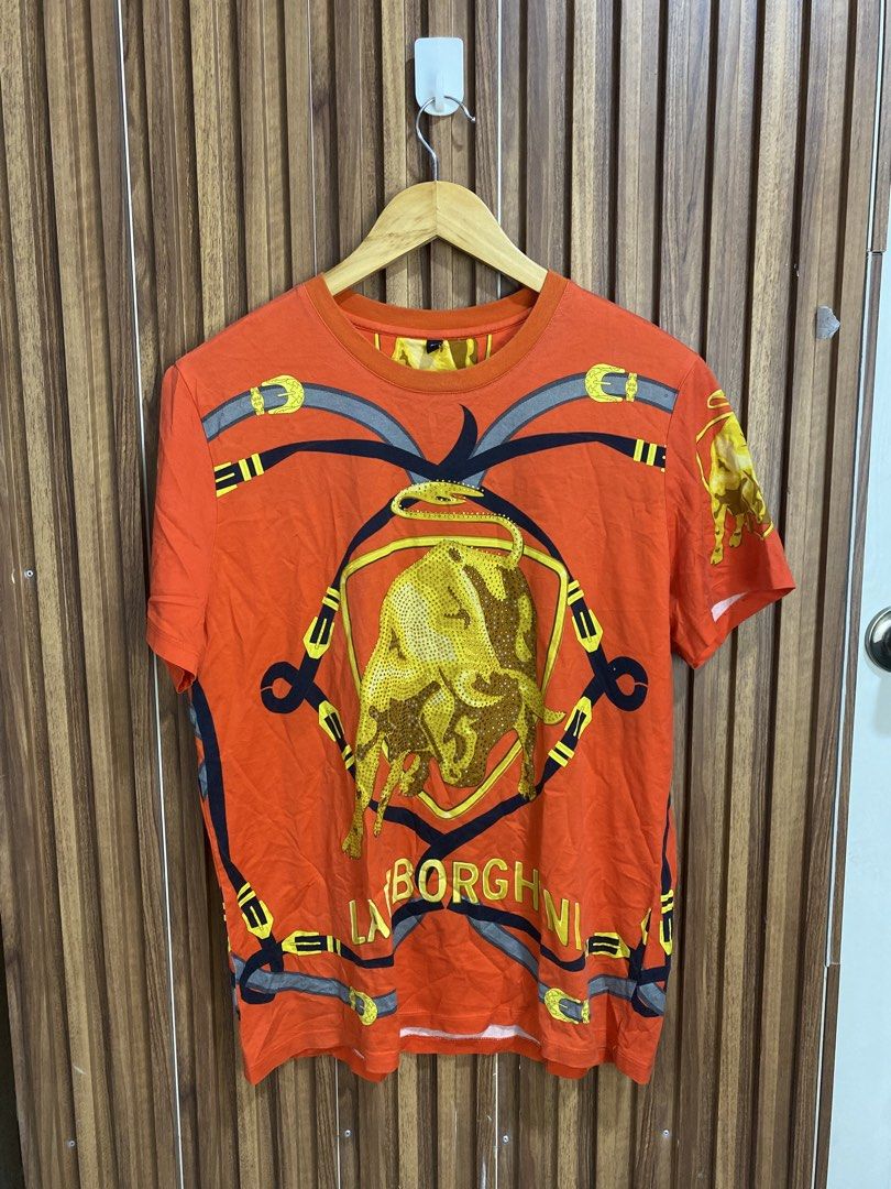Automobili Lamborghini Graphic Tee Orange (Category: Tshirts & Polo Shirts ) located in Caloocan City. Submitted by heatedcaps4425 (Listing ID 1471098618, Image 2). Description: - Brand: Automobili Lamborghini - Color: Bright orange - Design: Bold gold rhinestone bull logo on front, silver bull shield on back - Details: All-over printed belt graphics, short sleeves, crew neck - Style: Casual streetwear - Condition: Excellent, well-maintained - Sh
