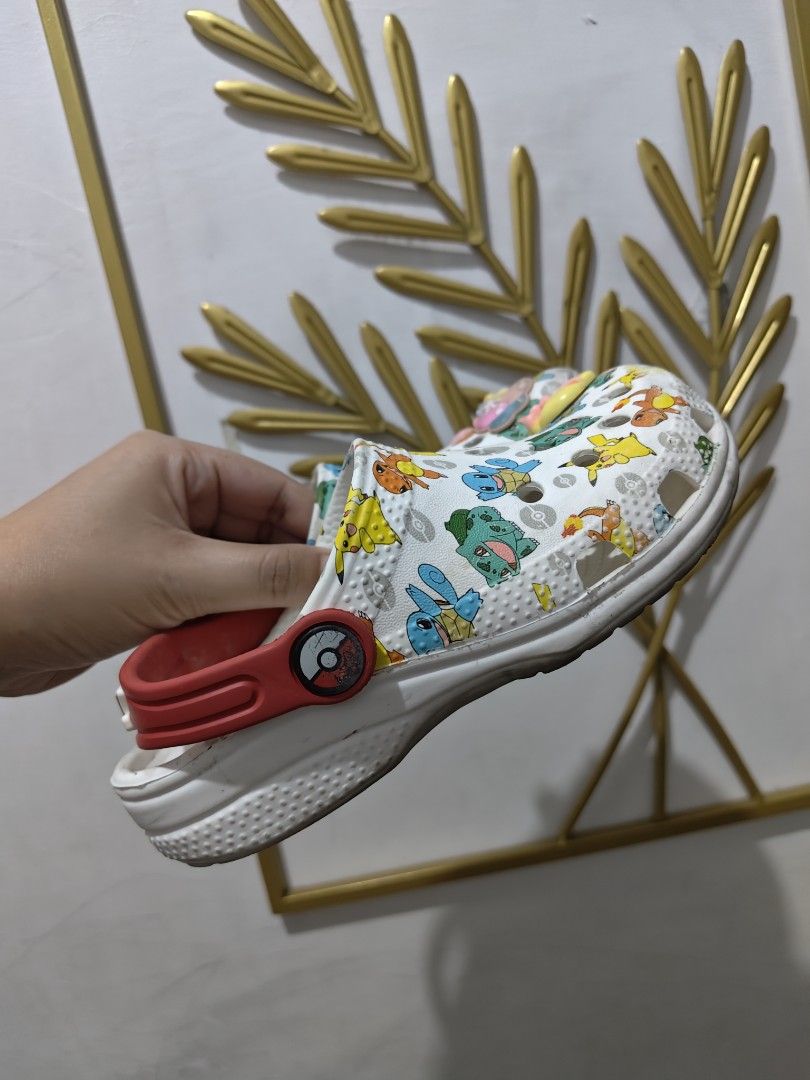 Crocs Pokemon Print Clogs with Red Straps and Jibbitz (Category: Babies & Kids Fashion) located in Mandaluyong City. Submitted by declutterpreloved2.0 (Listing ID 1471088146, Image 3). Description: - Design: White clogs covered in popular Pokemon characters (Pikachu, Squirtle, Bulbasaur, Charmander) - Details: Red heel straps featuring iconic Poke Ball pivot buttons - Accessories: Comes pre-decorated with cute colorful letter, bow, and flower cha