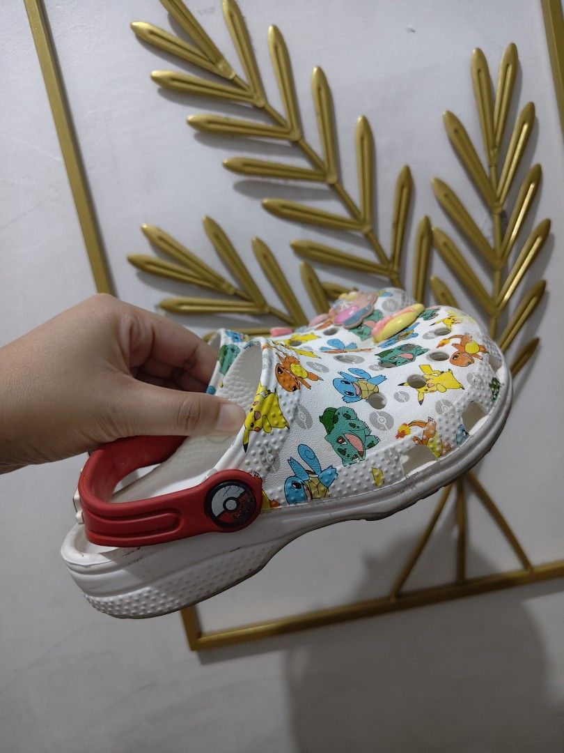 Crocs Pokemon Print Clogs with Red Straps and Jibbitz (Category: Babies & Kids Fashion) located in Mandaluyong City. Submitted by declutterpreloved2.0 (Listing ID 1471088146, Image 2). Description: - Design: White clogs covered in popular Pokemon characters (Pikachu, Squirtle, Bulbasaur, Charmander) - Details: Red heel straps featuring iconic Poke Ball pivot buttons - Accessories: Comes pre-decorated with cute colorful letter, bow, and flower cha