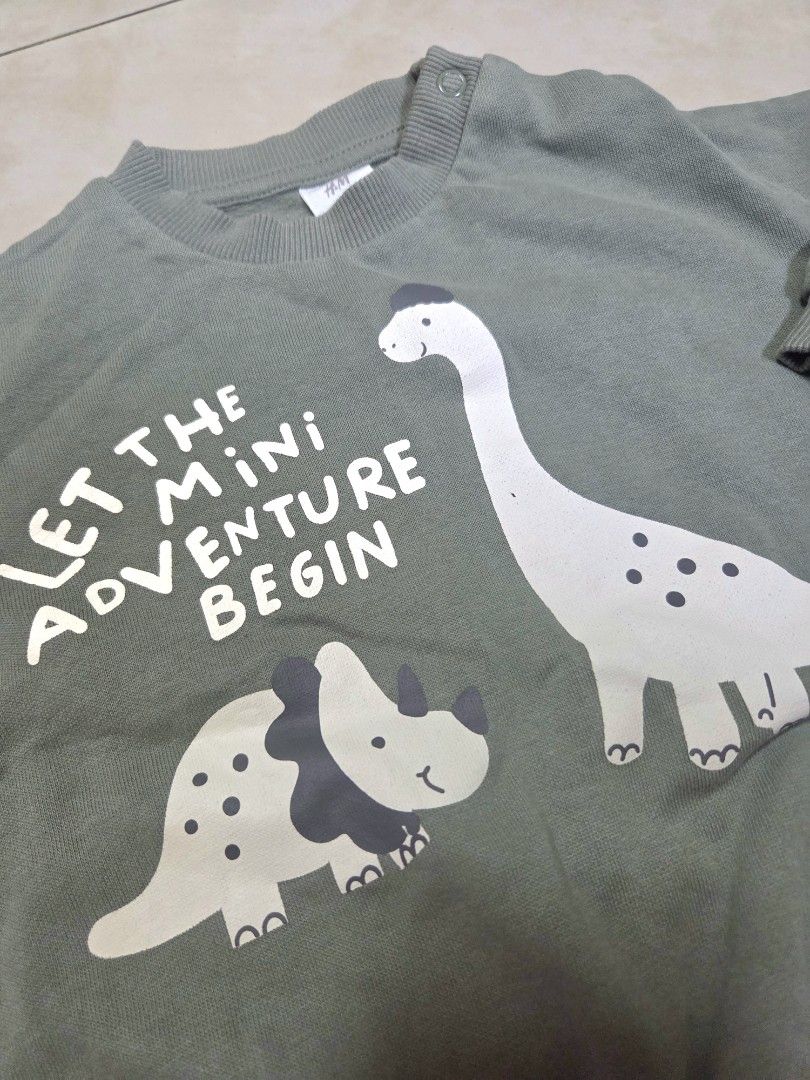 H&M Green Dinosaur Sweatshirt 6-9M (Category: Babies & Kids Fashion) located in Manila. Submitted by allthings.wellloved (Listing ID 1470963763, Image 2). Description: - Brand: H&M - Design: Sage green with cute dinosaurs and "Let the mini adventure begin" text - Size: EUR 74 / US 9M - Features: Convenient shoulder snap buttons for easy dressing, ribbed neckline, cuffs, and hem - Material: Super soft cotton blend inside and out - Condition: Excel