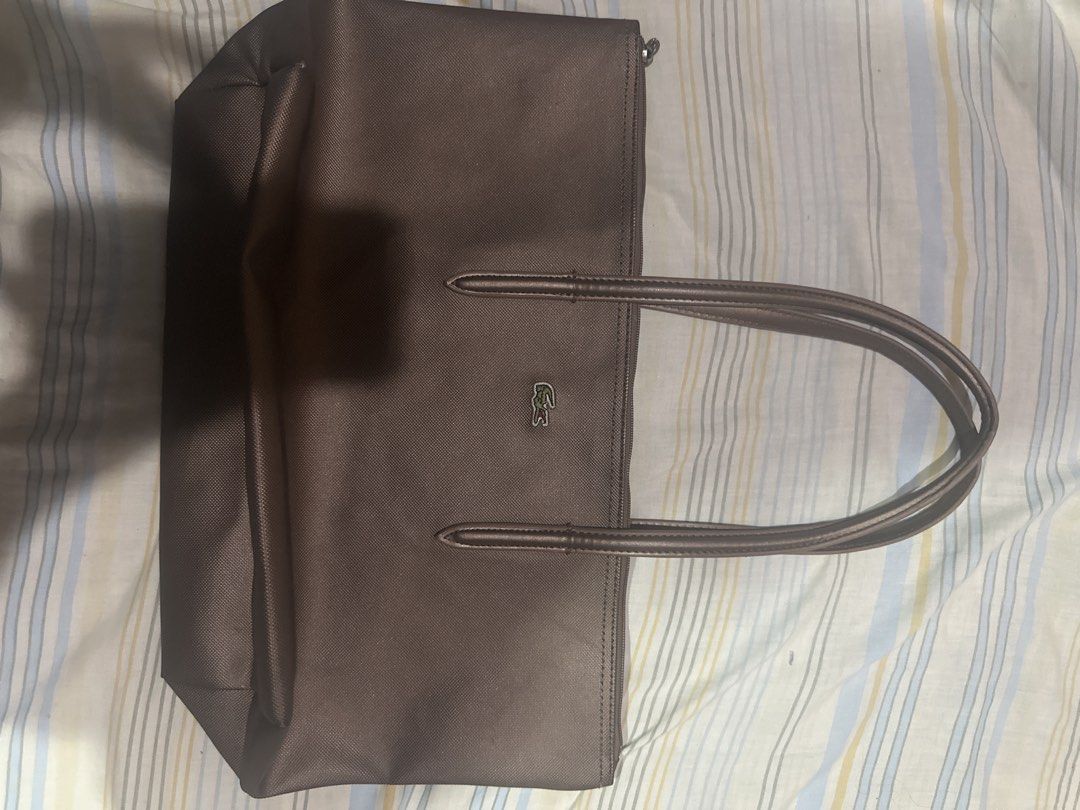 Lacoste Brown Tote Bag (Category: Tote Bags) located in Talisay. Submitted by kuyav (Listing ID 1471036583, Image 1). Description: - Brand: Lacoste - Color: Classic Brown - Features: Double shoulder straps, secure zip-top closure, structured canvas-like texture - Design: Iconic green crocodile logo on the front - Perfect for: Daily commuting, running errands, and everyday casual outfits -Issue:Leather strap has a tear, uses keychain for the zippe