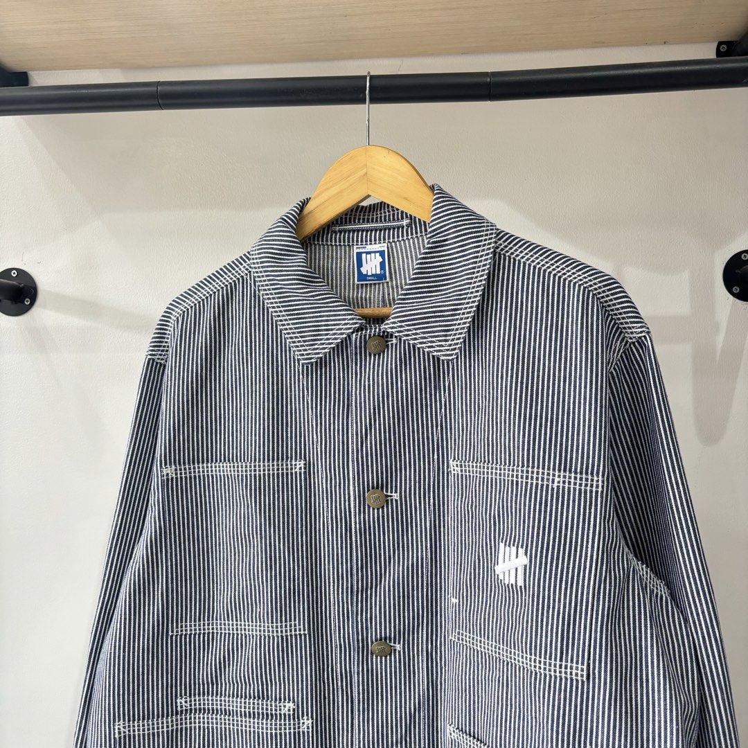 Undefeated Hickory Stripe Chore Jacket (Category: Coats, Jackets and Outerwear) located in Bambang. Submitted by heats777 (Listing ID 1471039458, Image 1). Description: - Brand: Undefeated - Item: Hickory Stripe Chore Jacket - Color: Blue and white - Size: Small (Regular fit) - Material: Polyester, cotton, and rayon blend - Features: Button closure, multiple front utility pockets, and embroidered logo near the lower left pocket - Condition: Excel