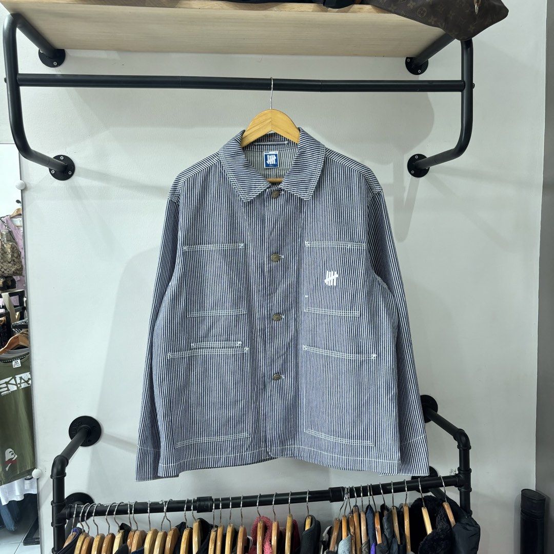 Undefeated Hickory Stripe Chore Jacket (Category: Coats, Jackets and Outerwear) located in Bambang. Submitted by heats777 (Listing ID 1471039458, Image 2). Description: - Brand: Undefeated - Item: Hickory Stripe Chore Jacket - Color: Blue and white - Size: Small (Regular fit) - Material: Polyester, cotton, and rayon blend - Features: Button closure, multiple front utility pockets, and embroidered logo near the lower left pocket - Condition: Excel