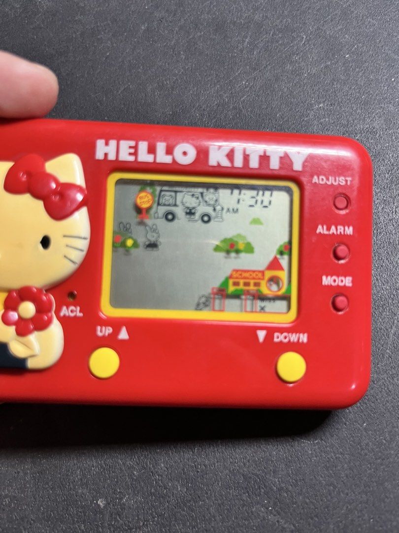 Vintage Hello Kitty Tomy LCD Game Electronic Handheld (Category: Interactive Toys) located in Caloocan City. Submitted by snapshop_ph (Listing ID 1471018923, Image 3). Description: Red handheld LCD game from Sanrio and Tomy. Kitty character molded on the left side holding a flower. Digital screen powers on, displays clock and game graphics like a school building and bus stop. Yellow control buttons below the screen. Battery cover on back, origina
